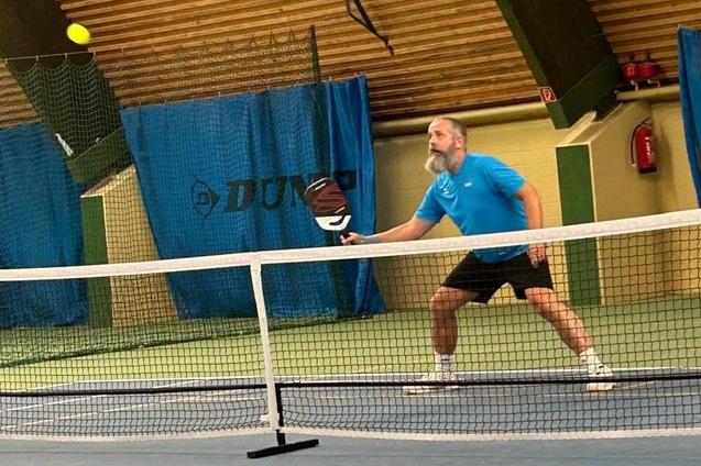 Mann spielt Tennis in einer Halle, schwingt den Schl&auml;ger, w&auml;hrend ein gelber Ball in der Luft ist.