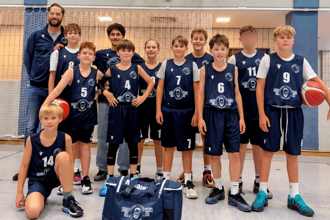 Gruppe von zw&ouml;lf Jugendspielern in Basketballtrikots mit einem Trainer in einer Sporthalle vor einem Korb.