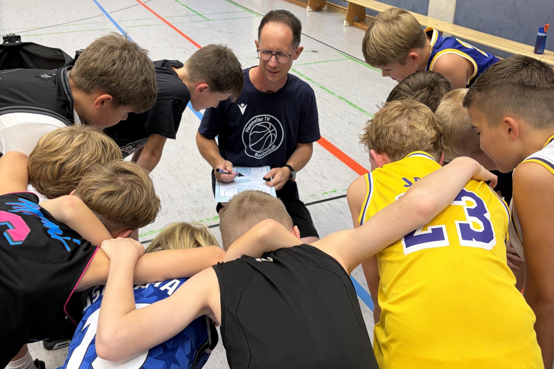 Trainer erkl&auml;rt Spielz&uuml;ge einer Basketballmannschaft, umringt von aufmerksamen Jugendspielern in Sportkleidung.