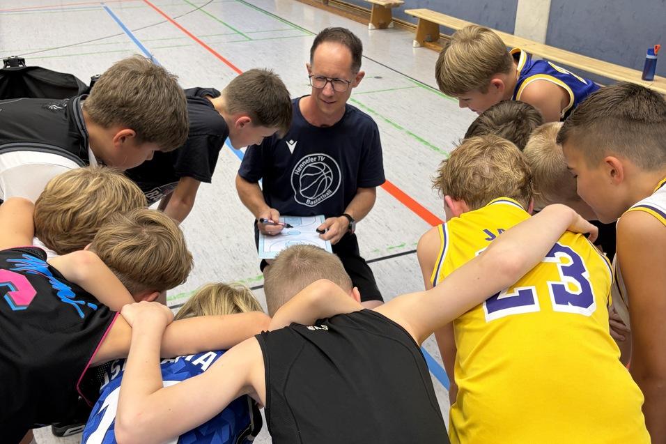 Trainer erkl&auml;rt Spielz&uuml;ge einer Basketballmannschaft, umringt von aufmerksamen Jugendspielern in Sportkleidung.