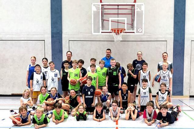 Gruppenzusammenkunft von Kindern und Trainern in Basketballtrikots, umgeben von Basketballs, in einer Turnhalle.