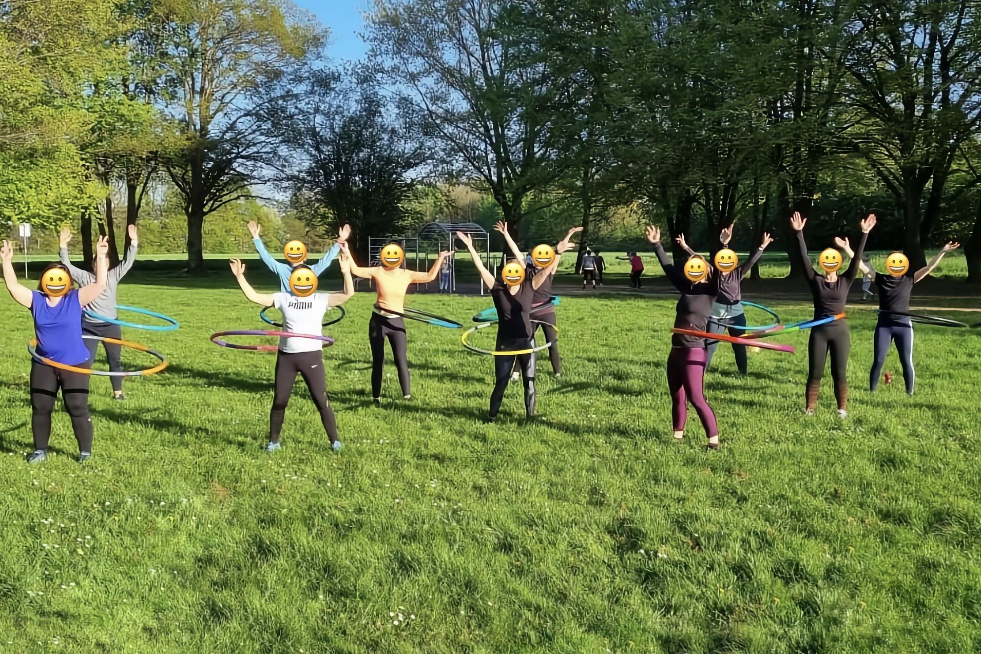 Gruppe von Personen mit Emoji-Gesichtsmasken, die fröhlich Hula-Hoops in einem Park nutzen.