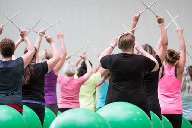 Gruppentraining mit Frauen, die auf Fitnessb&auml;llen sitzen und mit St&ouml;cken im Takt bewegen.