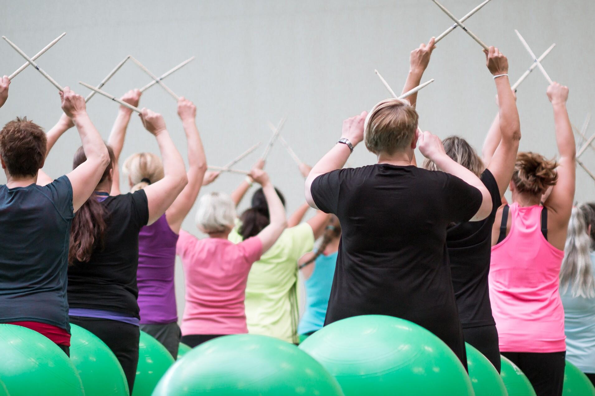 Gruppentraining mit Frauen, die auf Fitnessbällen sitzen und mit Stöcken im Takt bewegen.
