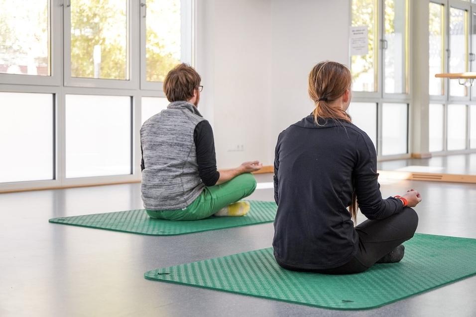 Zwei Personen sitzen meditierend auf gr&uuml;nen Yogamatten in einem hellen Raum mit gro&szlig;en Fenstern.