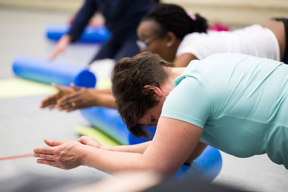 Mehrere Personen üben eine Sportübung auf blauen Yoga-Rollen in einem Kursraum für Fitness oder Rehabilitation.