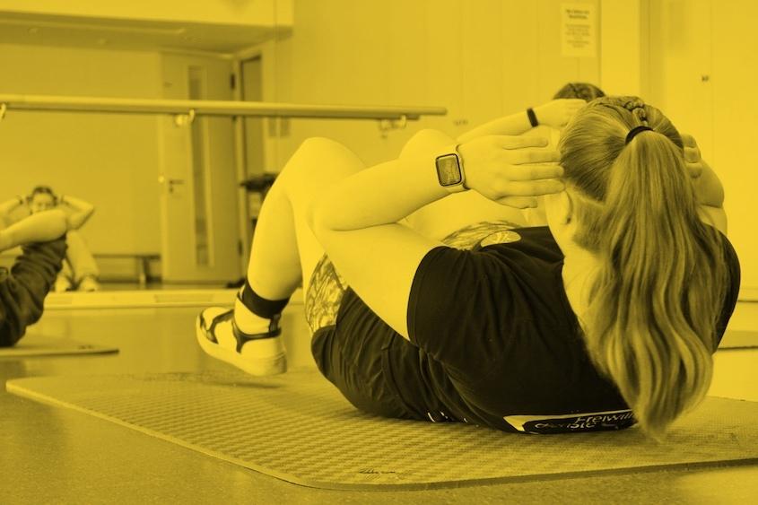 Frau macht Sit-ups auf einer Fitnessmatte in einem Raum mit Spiegeln und weiteren Trainierenden im Hintergrund.