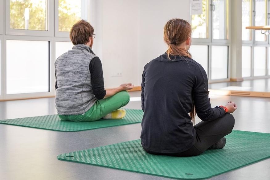 Zwei Personen sitzen meditierend auf grünen Yogamatten in einem hellen Raum mit großen Fenstern.