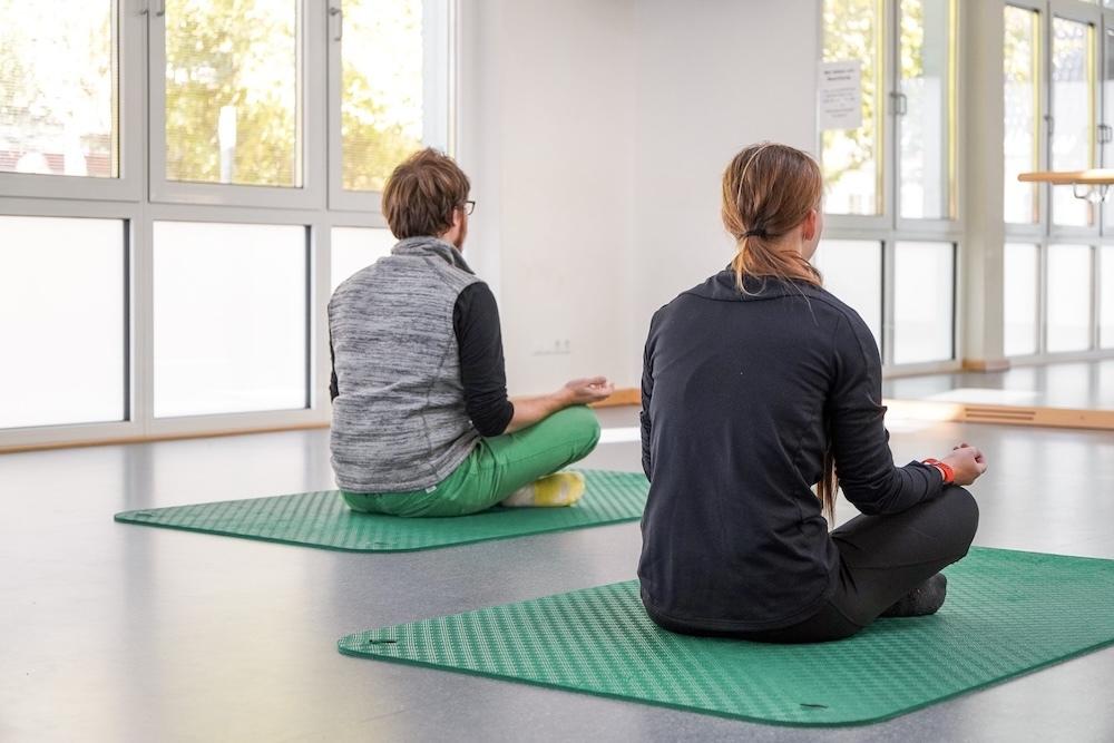 Zwei Personen sitzen meditierend auf grünen Yogamatten in einem hellen Raum mit großen Fenstern.