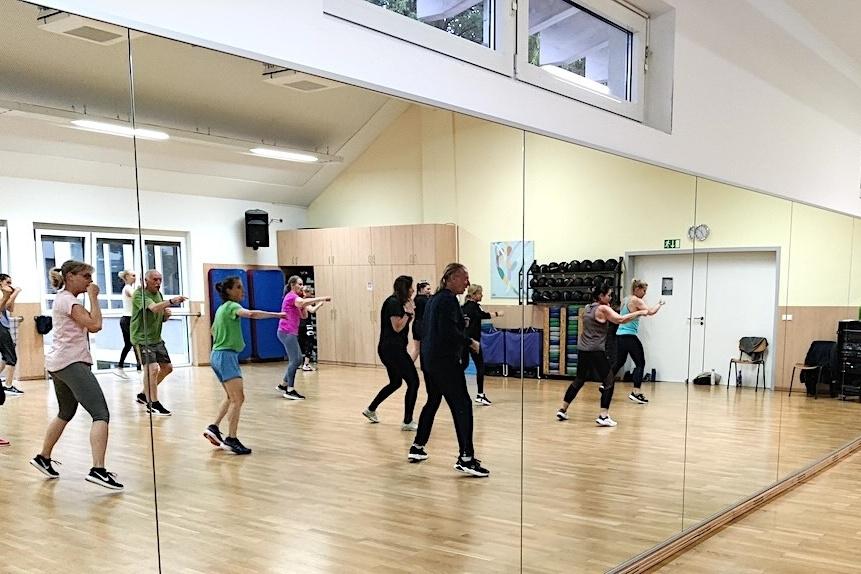 Gruppe von Menschen übt synchron in einem Tanzraum mit Spiegeln und Holzboden, ausgestattet mit Fitnessgeräten.