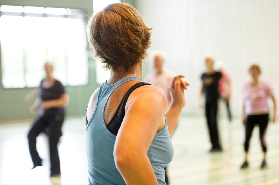 Eine Frau in Sportbekleidung zeigt eine Übung in einer Fitnessgruppe in einer Halle. Im Hintergrund sind weitere Teilnehmer.