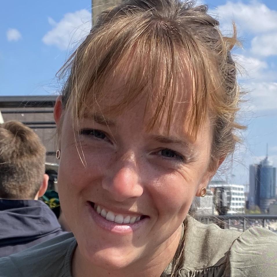 Lächelnde Frau mit braunen Haaren und einem grünen Oberteil, vor urbaner Kulisse unter blauem Himmel.