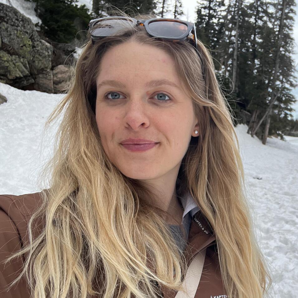 Frau mit langen Haaren und Sonnenbrille steht auf Schnee, im Hintergrund Bäume und Felsen.