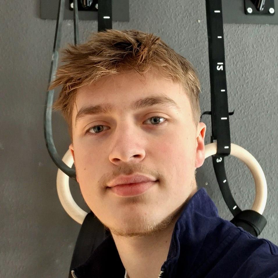 Junger Mann posiert vor einer Wandstation mit Ring- und Klimmzuggriffen in einem Fitnessraum.