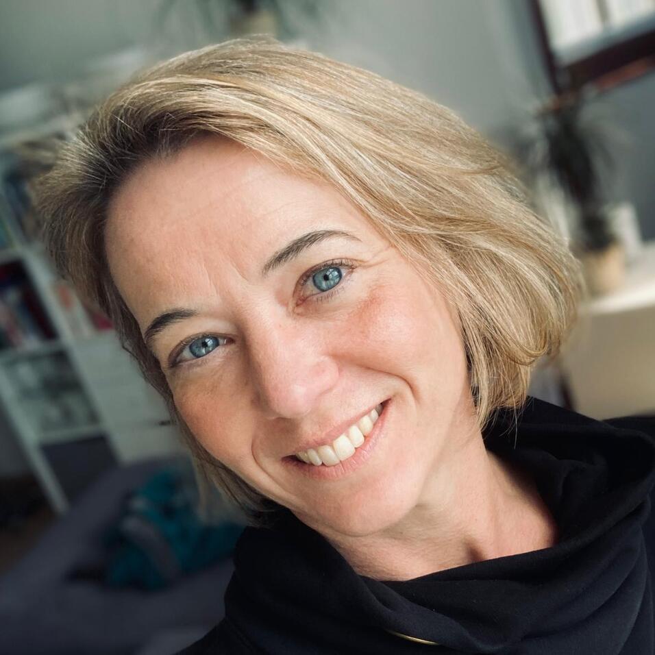 Porträt einer lächelnden Frau mit blonden Haaren, in einem gemütlichen Raum mit Pflanzen und Möbeln im Hintergrund.