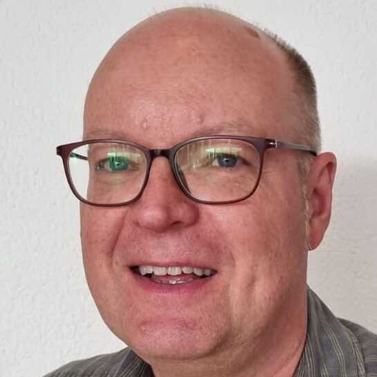 Porträt eines lächelnden Mannes mit Brille, kurz über dem Kopfhaar, vor einer hellen Wand.