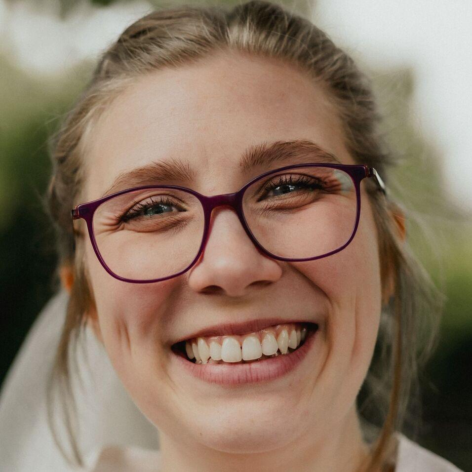 Lächelnde junge Frau mit Brille und Halbhochfrisur, Aufnahme im Freien mit verschwommenem Hintergrund.