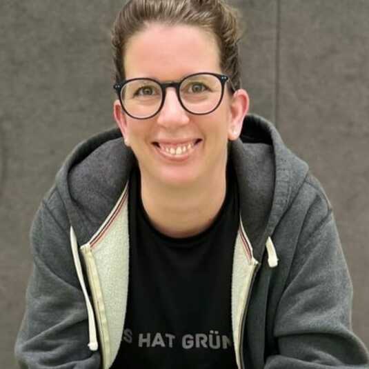 Frau mit Brille sitzt lächelnd auf einer Bank, trägt grauen Hoodie und schwarzes T-Shirt mit Aufschrift.