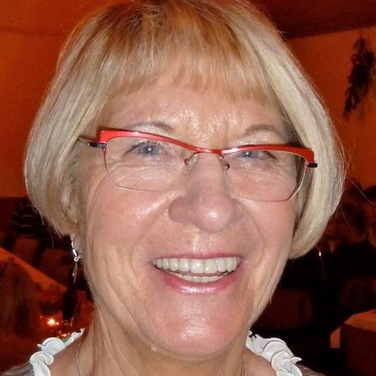 Ältere Frau mit blonden, kurzen Haaren und roten Brillenfassungen lächelt in einem gemütlichen Innenraum.