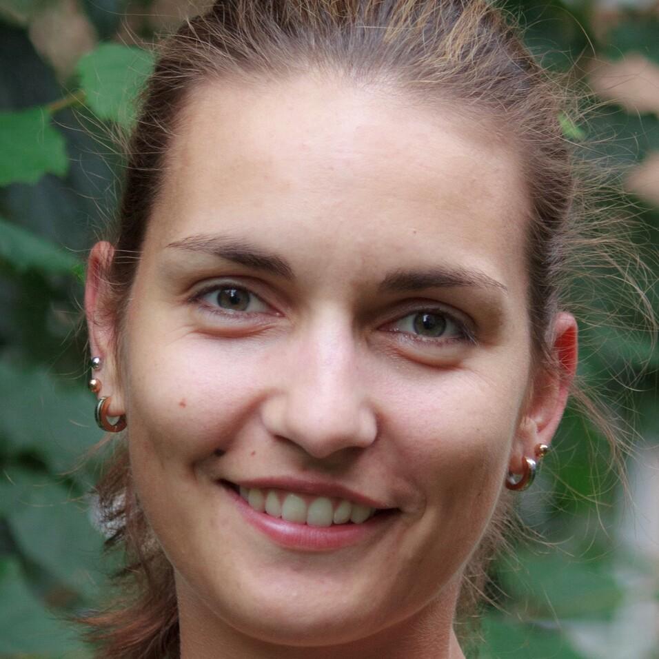 Porträt einer lächelnden Frau mit kurzen, hellbraunen Haaren und Ohrringen vor einem unscharfen grünen Hintergrund.