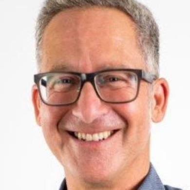 Portrait eines lächelnden Mannes mit Brille, grauem Haar und heller Bluse, vor weißem Hintergrund.