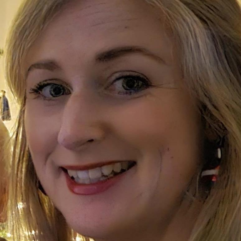 Nahaufnahme einer lächelnden Frau mit langen blonden Haaren und Ohrhängern vor einem sanften, warmen Hintergrund.