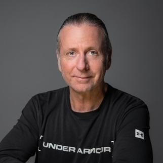Porträt eines Mannes mit kurzen, grauen Haaren, der in einem schwarzen Shirt mit Logo in neutralem Hintergrund sitzt.