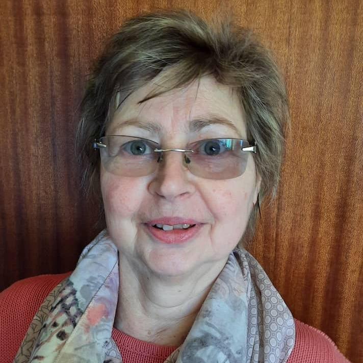 Lächelnde Frau mit Brille und farbigem Schal vor einer Holzmaserung.