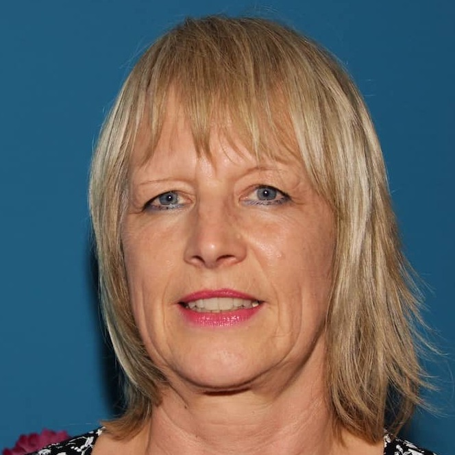 Portr&auml;t einer Frau mit blonden, schulterlangen Haaren, die ein schwarzes Bl&uuml;mchenmuster tr&auml;gt, vor blauem Hintergrund.