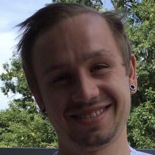 Ein lächelnder junger Mann mit kurzen Haaren und Ohrsteckern steht vor einer grünen Landschaft.