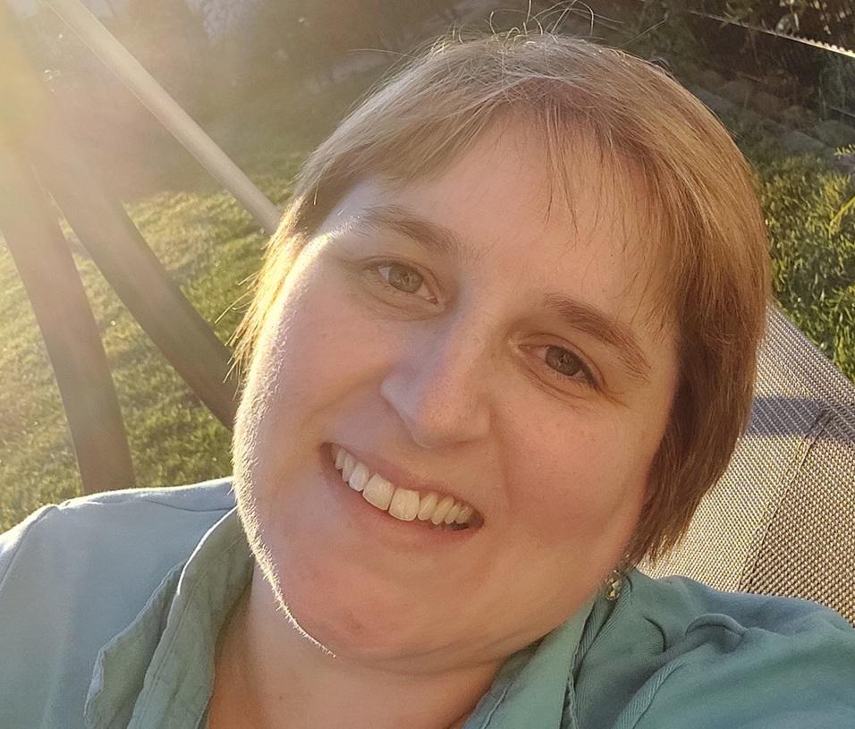 Frau mit kurzer Frisur lächelt freundlich in die Kamera, im Hintergrund Sonne und Grünflächen sichtbar.