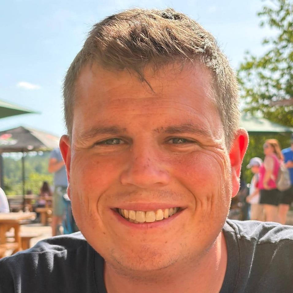 Lächelnder Mann mit kurzer Frisur sitzt im Freien, Hintergrund mit unscharfen Menschen und Sonnenschein.