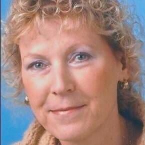 Portrait einer Frau mit lockigem, blondem Haar vor blauem Hintergrund, sanft lächelnd in heller Kleidung.