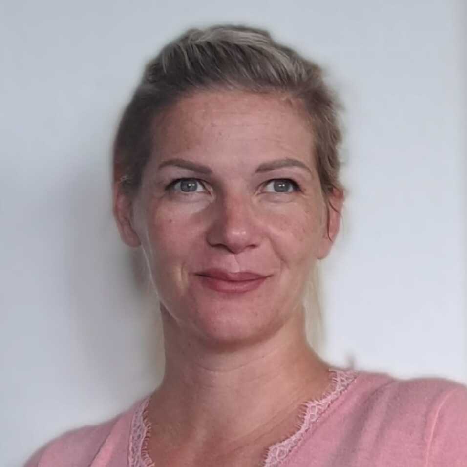 Porträt einer lächelnden Frau mit blonden Haaren, im rosa Shirt, vor einem hellen Hintergrund.
