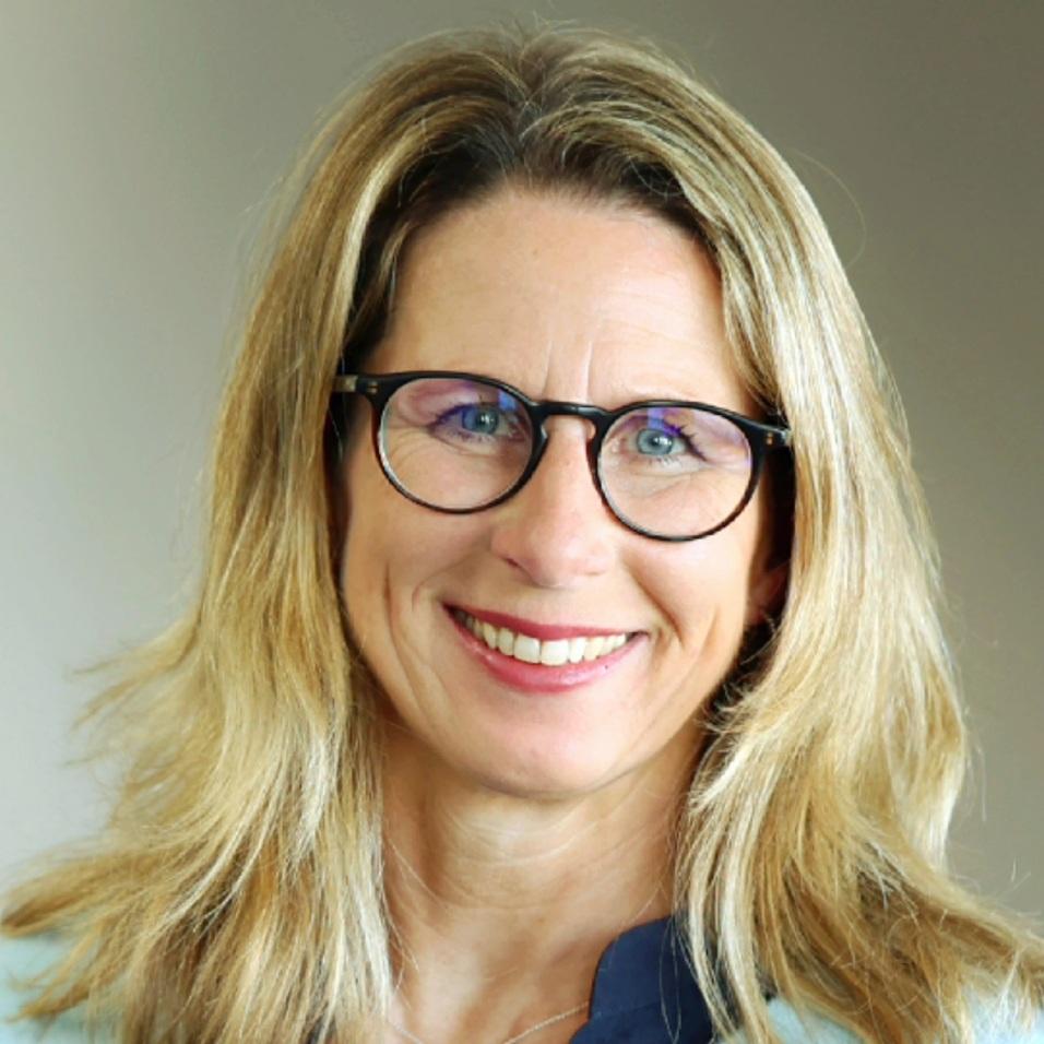 Porträt einer lächelnden Frau mit blonden Haaren, Brille und hellblauem Pullover vor neutralem Hintergrund.