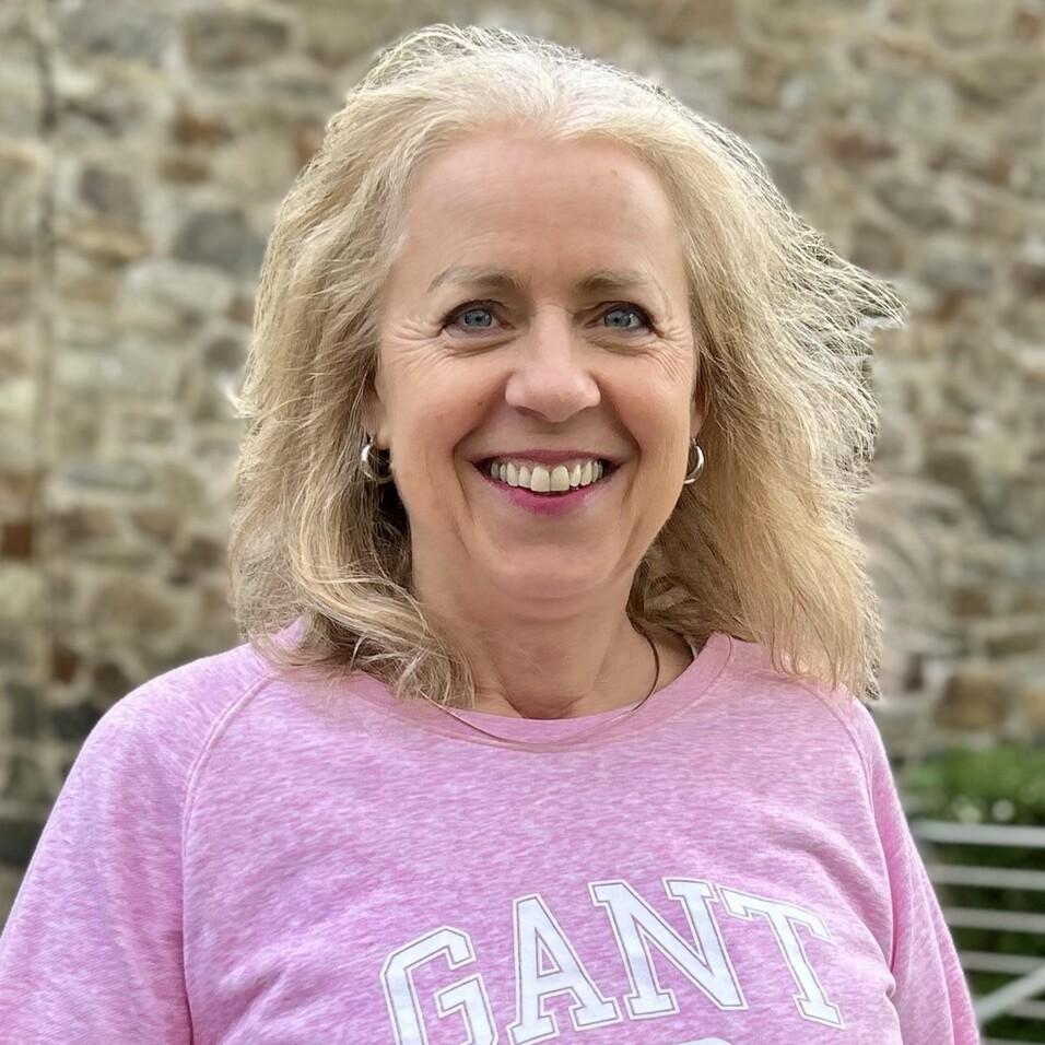 Lächelnde Frau mit blonden, lockigen Haaren trägt ein rosa Sweatshirt vor einer steinernen Mauer.