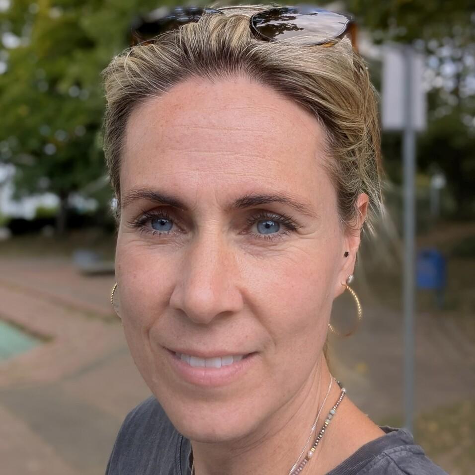 Porträt einer Frau mit blonden Haaren und Ohrringen, die lächelt, vor einem parkähnlichen Hintergrund.