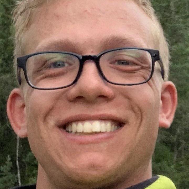 Lächelnder junger Mann mit Brille in grüner Jacke vor einer bewaldeten Umgebung.