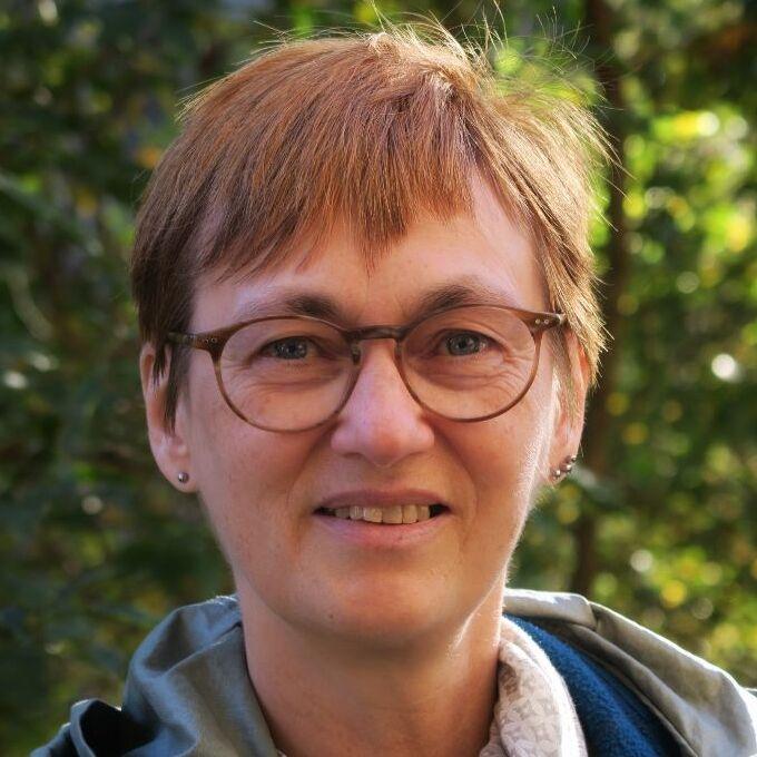 Porträt einer Frau mit kurzen, braunen Haaren und Brille, lächelnd vor unscharfem, grünem Hintergrund.