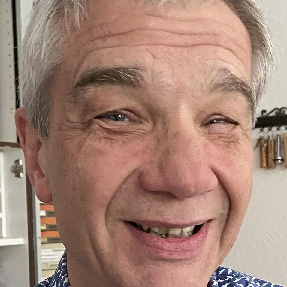 Lächelnder Mann mit grauen Haaren und blauem Hemd mit Vogelmotiven, Hintergrund mit Regalen und Werkzeug.