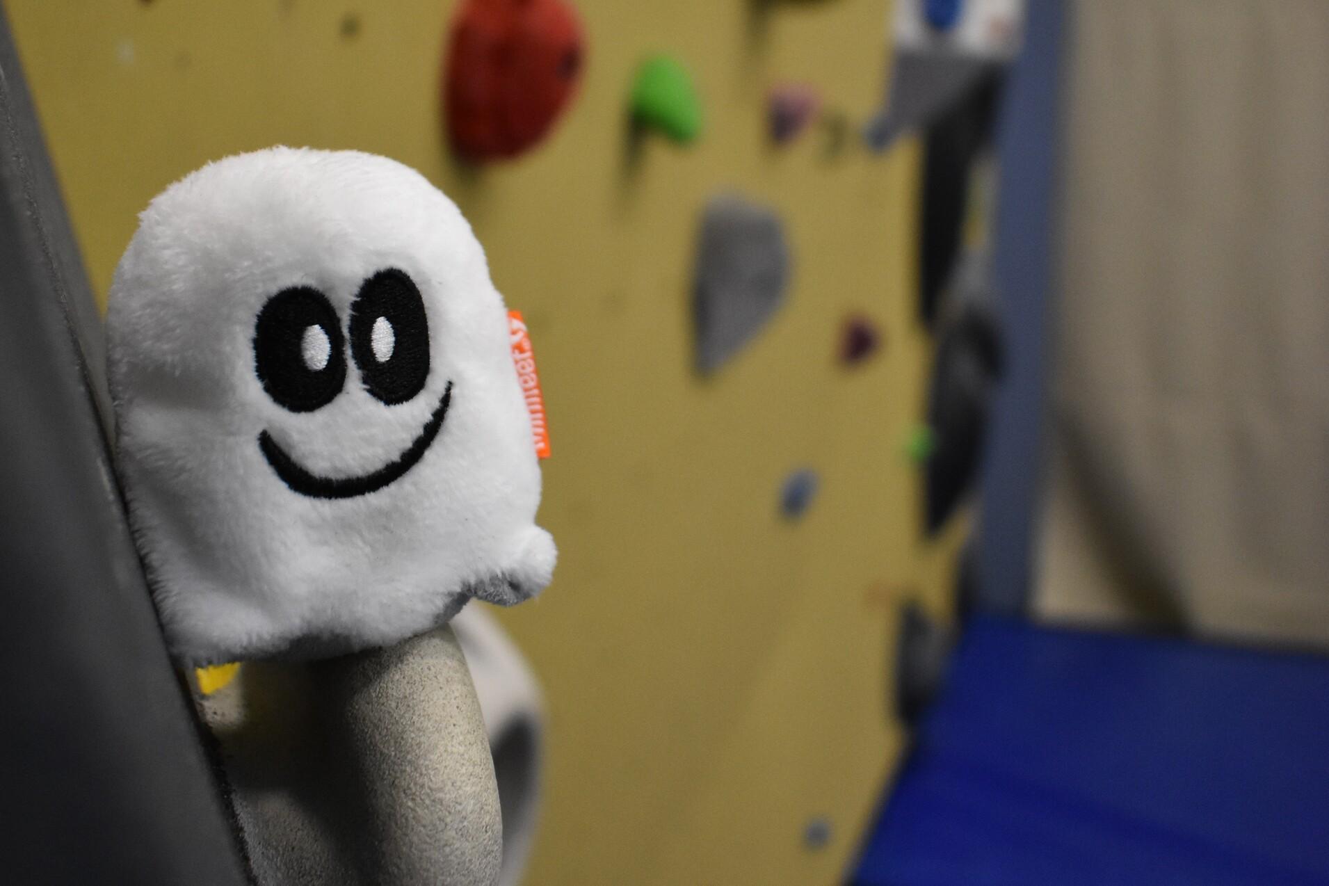 Pl&uuml;schgespenst mit l&auml;chelndem Gesicht an einer Kletterwand mit bunten Griffen im Hintergrund.
