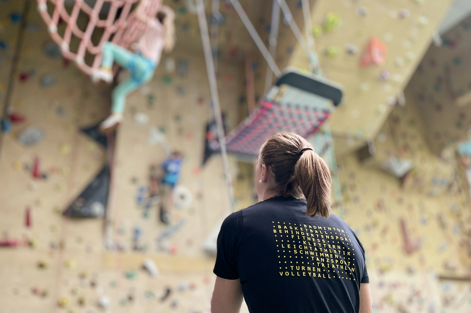 Eine Klettererin schaut auf eine Kletterwand mit verschiedenen Routen und einer Netzschaukel im Hintergrund.