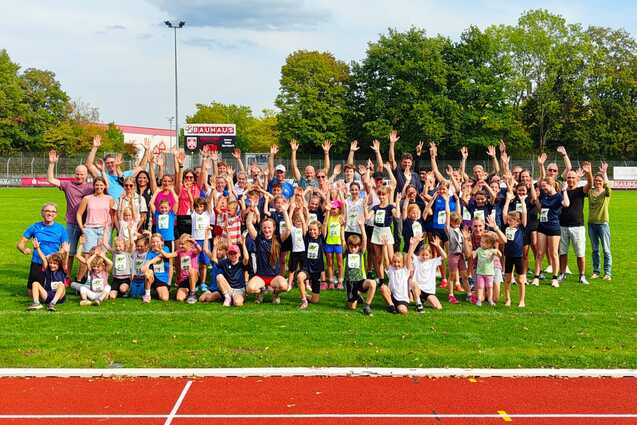Gruppenfoto von Kindern und Erwachsenen mit erhobenen H&auml;nden auf einer Sportanlage, gr&uuml;ne Wiese im Hintergrund.