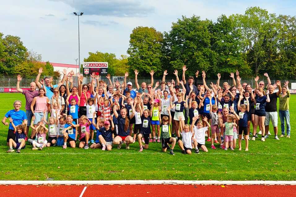 Gruppenfoto von Kindern und Erwachsenen mit erhobenen Händen auf einer Sportanlage, grüne Wiese im Hintergrund.