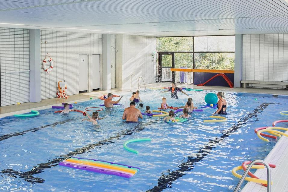 Innenbereich eines Schwimmbades mit fröhlichen Schwimmern, Schwimminstrumenten und bunten Aufblasspielzeugen im Wasser.