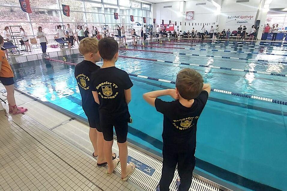 Drei Kinder in schwarzen T-Shirts stehen am Beckenrand eines Schwimmbeckens und beobachten die Wettk&auml;mpfe.
