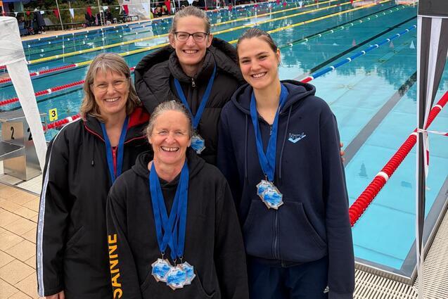 Vier Schwimmerinnen mit Medaillen stehen vor einem Schwimmbecken, umgeben von Zelten und B&auml;umen an einem grauen Tag.