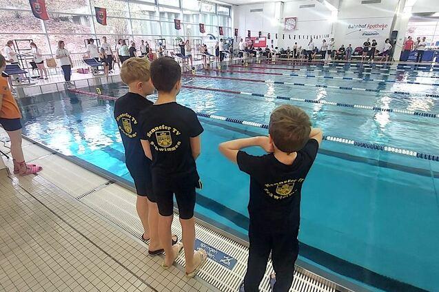 Drei Kinder in schwarzen T-Shirts stehen am Beckenrand eines Schwimmbeckens und beobachten die Wettk&auml;mpfe.