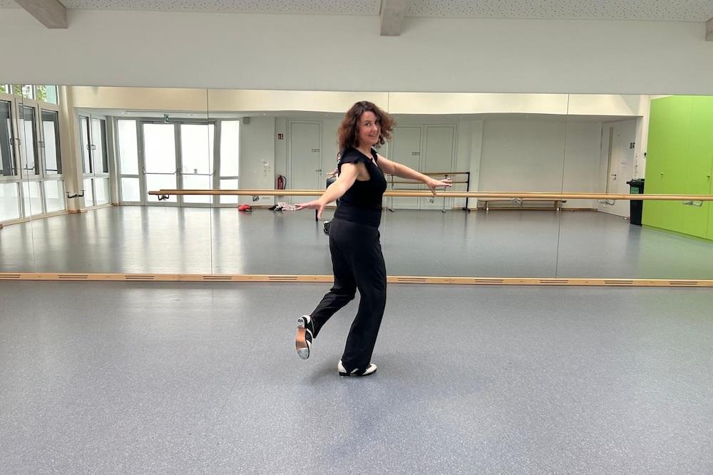 T&auml;nzerin in schwarzer Kleidung f&uuml;hrt eine Bewegung im Tanzstudio vor einem gro&szlig;en Spiegel aus.