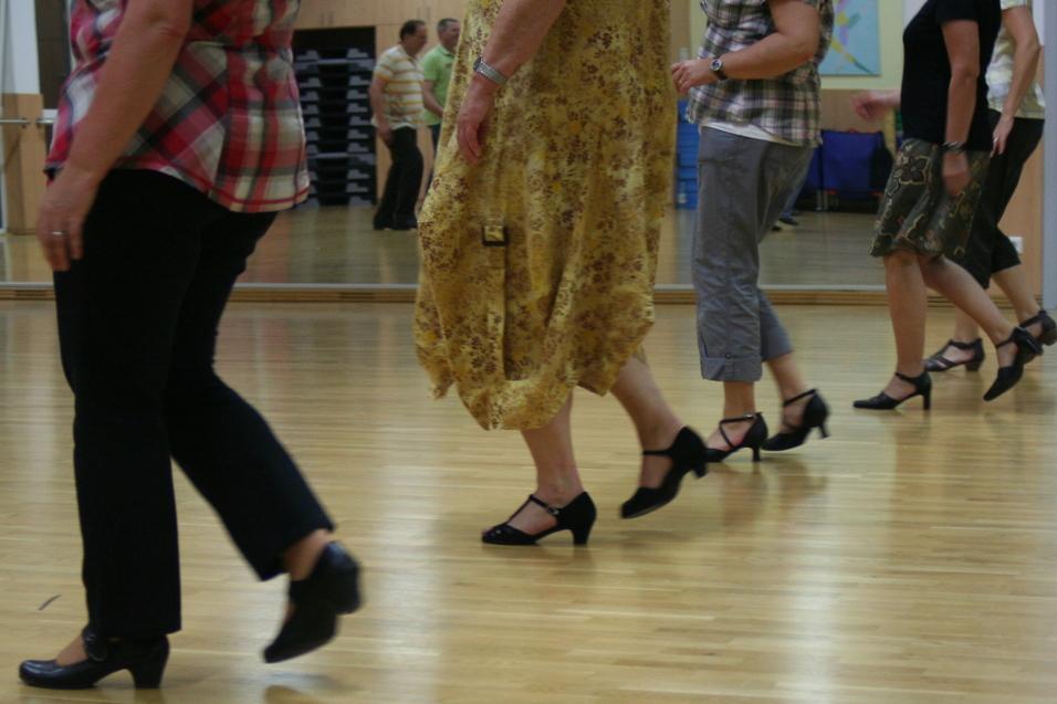 Tanzende Frauen in verschiedenen Outfits und Schuhen auf einem hellen Parkettboden in einem Tanzraum.